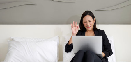 Asian woman in black suit use the laptop computer to work in the morning in bed. Waved her hand over...