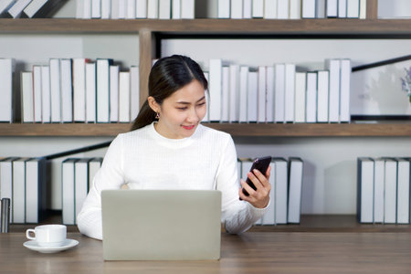 Young asian woman reading text message on mobile phone while doing her thesis on laptop computer in...