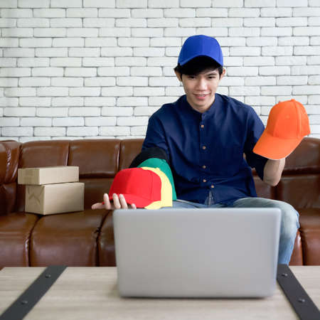 Young asian man sells various colors of cap hat online via application on laptop computer. Small home business concept.の写真素材