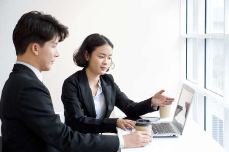 A salesperson in a suit presents the product through laptop computer to the customer. Young businessman listening attentively while holding a cup of coffee.の写真素材