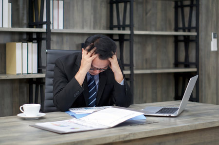 Sad Asian businessman holds his temple with both hands, sitting in the office. The concept of...