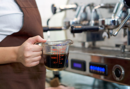 Barista dressed in brown apron holding a measuring cup fill with hot coffee. Morning atmosphere in a...