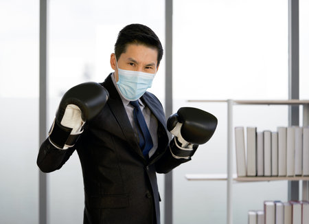Asian businessman dressed in suit and necktie, wearing a face mask and a boxing glove. Standing...