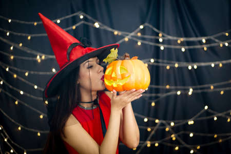 Caucasian woman in red dress  kissing pumpkin carved into a face stand in front of black curtain decoration with lighting at halloween party.の写真素材