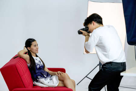 Young asian photographer is photographing a model posing on a red sofa for advertising in a magazine.の写真素材