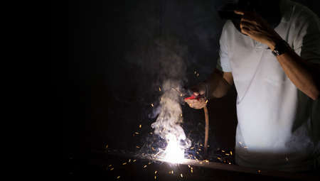 Metal industry worker in white t-shirt with collar welding sheet metal in a factory. Sparkler on dark background, close-up. Heavy work in factory.の写真素材