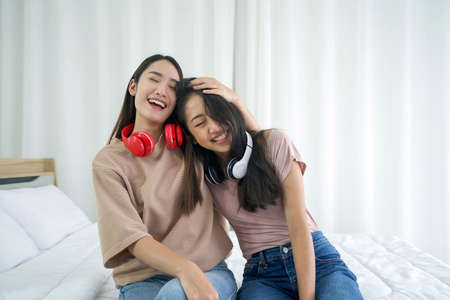 Young asian teen and her friend with headphones laughing happily while sitting in the bed to relax in the bedroom, enjoy leisure weekend at home. Stress free conceptの写真素材