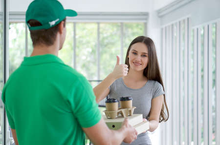 The delivery staff in a green uniform handed the pizza box and coffee cup to the young customer. The woman raised her thumbs satisfied with the food delivery service. Food and parcels delivery conceptの写真素材