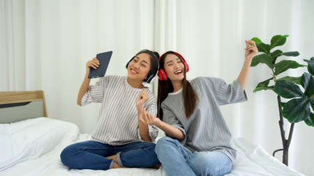 Young asian teen and her friend with headphones listening to music while sitting in the bed to relax in the bedroom, enjoy leisure weekend at home. Stress free conceptの写真素材