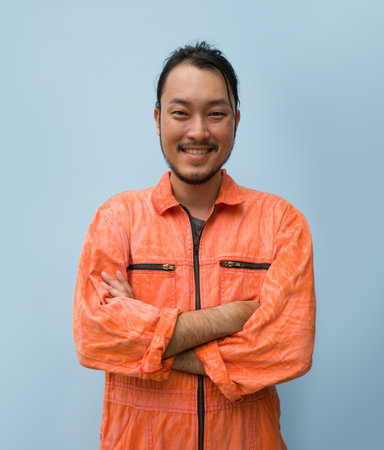 Young long hair asian man with mustache and beard stand smiling with his arms crossed. Portrait of industrial people on blue background.の写真素材