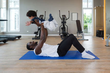 Young short curly black hair father  with moustache and beard in sportswear playing with his son after finish exercise routine on yoga mat. Cardio machines are on the background at the gym.の写真素材