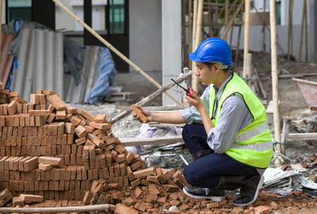 Asian contractor with safety helmet, reflective vest and rubber boots checking quality of brick while holding walkie talkie. Young engineer working in construction site.の写真素材