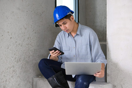 Young asian engineer in hardhat transfer data from mobile phone to laptop computer while sit on a...