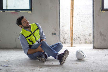 Young caucasian man in safety vest gripped his knee and screams in pain. Due to injuries at the construction site.の写真素材