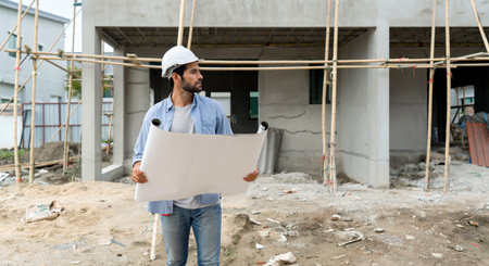 Young engineer in a construction helmet holding a floor plan while looking at the progress of real...