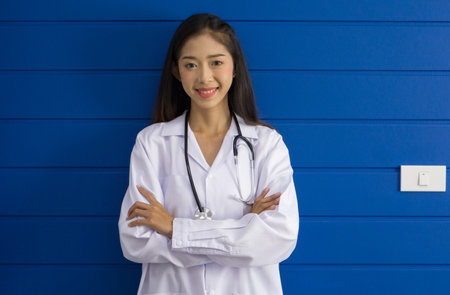 Young asian doctor in white gown and stethoscope stand with arm cross in front of blue wall.の写真素材