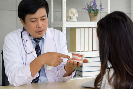 Asian dentist in white gown and stethoscope pointing finger at tooth model, explain to young patient about orthodontics and braces. Help reduce gum pain. Healthcare and medicine concept.の写真素材