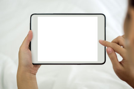 Back view of woman typing on blank screen tablet computer while sit relax on bed with white duvet....