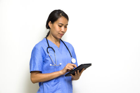 Woman physical therapist in blue uniform and stethoscope typing on tablet computer. Stand in front of white wall.の写真素材