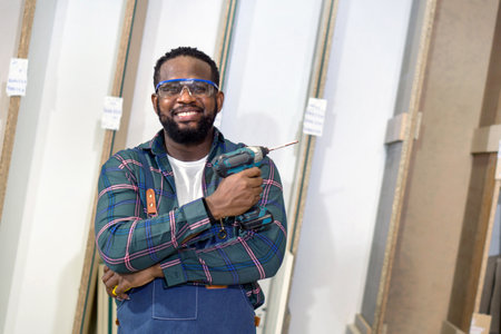 Short black hair man with moustache and beard stand with arm folded, holding cordless electric drill. Plywood storage compartment are on the background. Carpenter worker work in a furniture factory.の写真素材