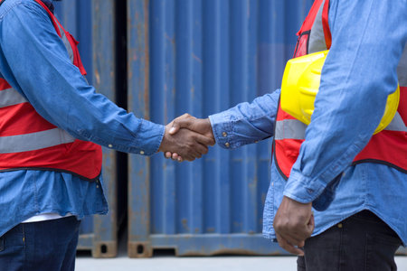 Two workers clad in safety helmets and reflective vests shake hands in front of a massive steel...