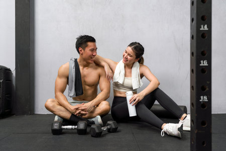 Young asian couple amidst workout on gym floor, surrounded by dumbbells, engaging in a moment of rest.の写真素材