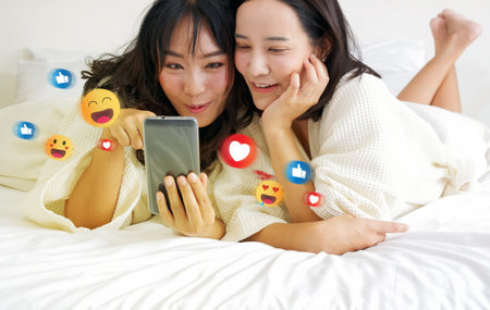 Two cheerful asian women, nestled in bed in bathrobes, are indulged in gleeful selfie-session,...