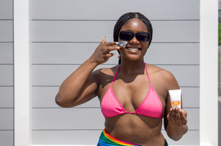 A dark-skinned lady applying sunscreen on her face, holding a sunscreen tube, dressed in summer attire, standing against a  wall.の写真素材