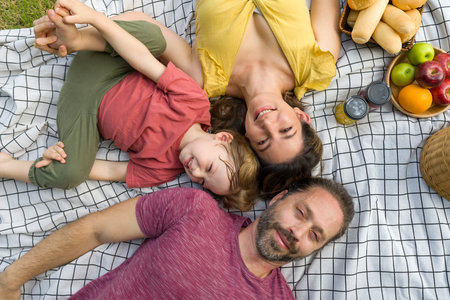A happy family is sprawled comfortably on a picnic blanket in a sunny park, enjoying food and each other's company.の写真素材