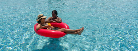 Two ladies in bikini are joyfully floating together on a big inflatable ring in blue water.