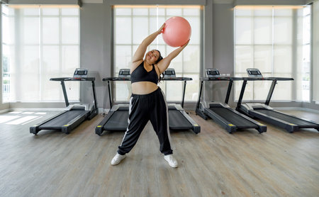 A woman is in a gym, gripping a bright pink ball with both hands, ready to exercise.の写真素材