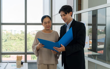 A collaborative work effort, a discussion on business report. Young male in suit and senior female looking at document inside a blue folder. They are standing in a well-lit office with a large windowの写真素材