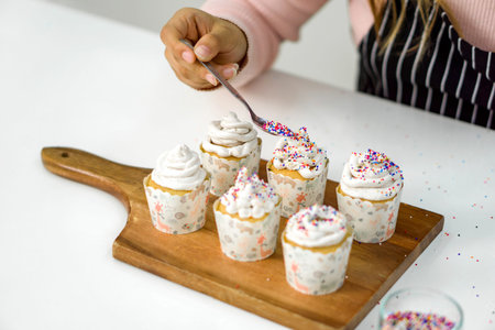 Closeup woman hand holding spoon with multicolor frosting, decorating cupcake on a wooden board,...