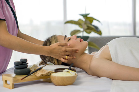 Beautiful asian woman getting face massage in beauty spa. Young woman with closed eyes relaxing in indoor spa while getting head massage. Serene woman relaxing  in a beauty center. Side view.の写真素材