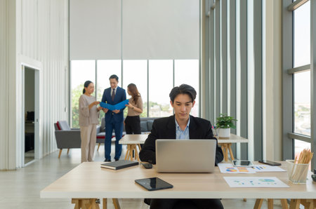 Young businessman sit in the foreground, working on a laptop. In the background, three other individuals are engaged in a conversation, sharing a document. Atmosphere in office with large window.の写真素材