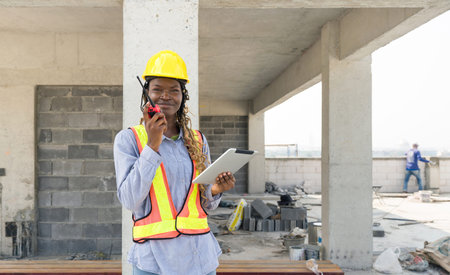 A builder in safety uniform standing at a building site, holding a walkie-talkie communicating with...