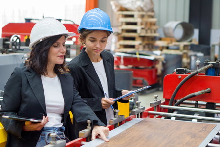 Two women dressed in black suit and safety helmet are inspecting work in a factory setting.の写真素材
