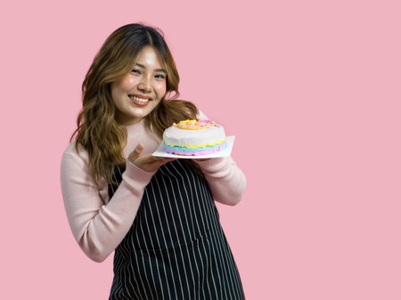 Young asian woman with an apron in celebratory or festive mood holding colorful cake with various sprinkles on the topping. Portrait on pink background with studio light.の写真素材