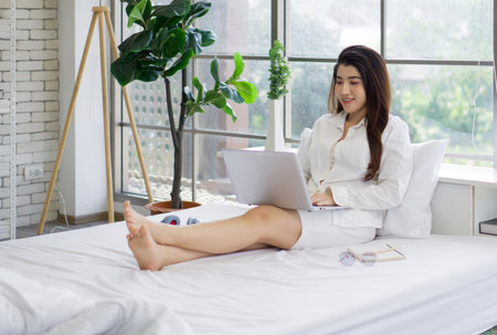 Young asian woman is working on a laptop computer while sitting comfortably on a white bed. Work from home concept.の写真素材