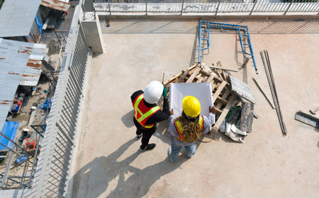 Two worker in helmet are busy, looking at blueprint on a sunny day at a construction site. High angle view.の写真素材