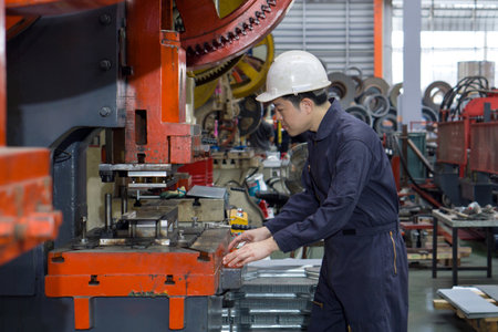 A man in a hardhat and mechanic coverall dust protection suit using a machine to shape metal parts safely. Atmosphere in manufactory with large machinery.の写真素材