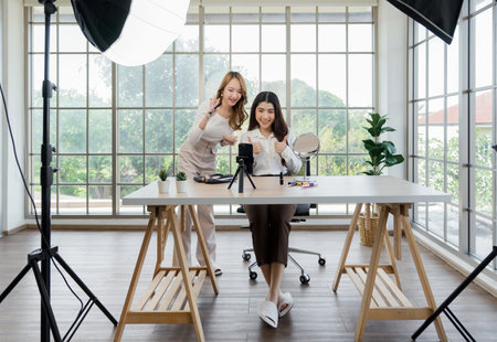 Two asian women creating content for social media. They are using a studio setup that include a smartphone mounted on a tripod, lighting equipment and various makeup products arrayed on the table.の写真素材