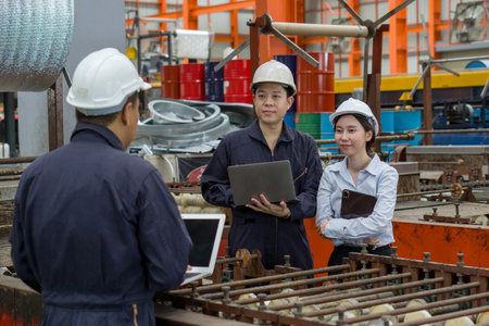 Three workers in a factory discuss a project using laptop computer. Everyone wearing protective hardhat. Atmosphere in manufactory with large machinery.の写真素材