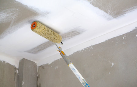 A paint roller is being used to apply a fresh coat of white paint on the ceiling of a newly renovated room. The wall have a smooth finish, indicating recent update.の写真素材