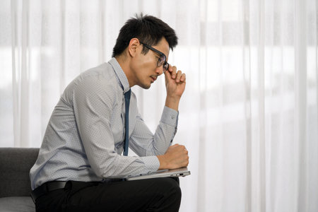 A man sit thoughtfully on a sofa, wearing a shirt and tie. He lightly touches his glasses, deep in...