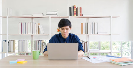 A person is concentrating on a laptop at a tidy wooden desk surrounded by bookshelves. The bright...