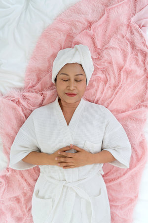 Senior asian woman dressed in a cozy white robe lies peacefully on a soft pink blanket with her hand...