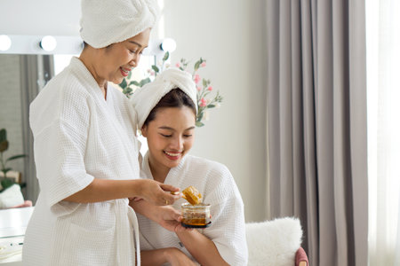 Asian mother and daughter share a joyful moment as they apply skincare product in a cozy home spa. They are dressed in comfortable robe and towel, surrounded by a tranquil atmosphere.の写真素材