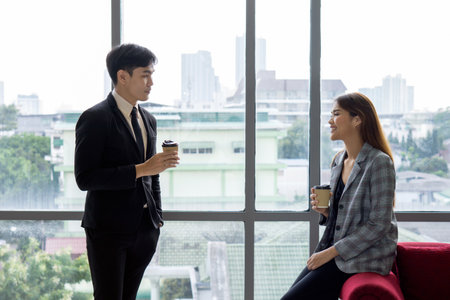 Professional stand conversing in a stylish office space, sipping coffee. The large window showcase a bustling city skyline, and bright daylight fill the room.の写真素材