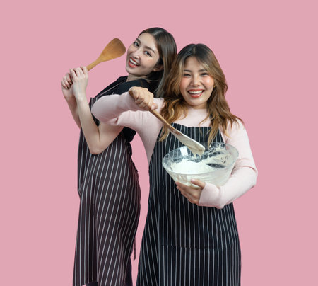 Two women are having fun baking together. They are wearing apron and smiling while mixing ingredients with a wooden spoon. Portrait on pink background with studio light. Isolatedの写真素材
